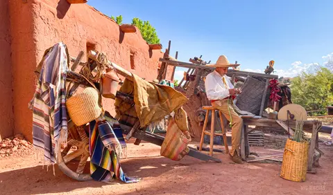 A man wearing a straw hat sits on a stool outside near a rustic wooden cart and adobe wall.