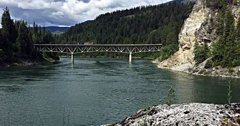 A bridge spans a wide river surrounded by forested hills and rocky banks.