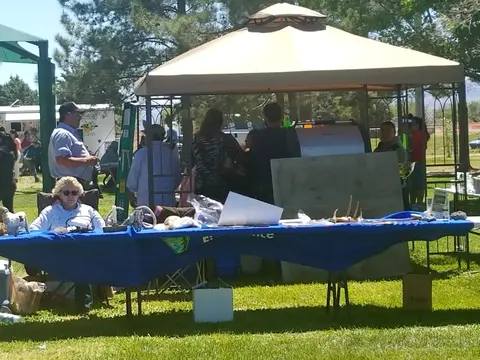 People gather around a table with a blue cloth under a canopy tent outdoors.