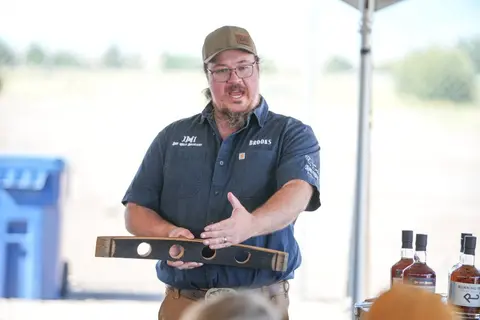 A man holding a wooden barrel stave and gesturing while speaking outdoors.