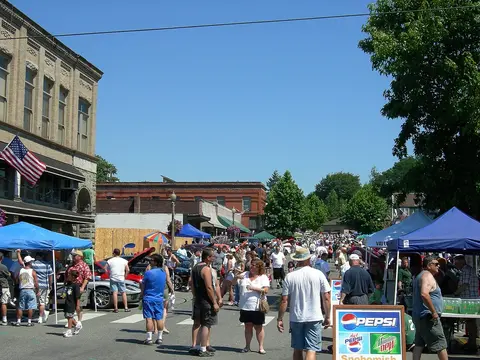 A street scene with a crowd of people walking among booths and parked cars under a clear blue sky.