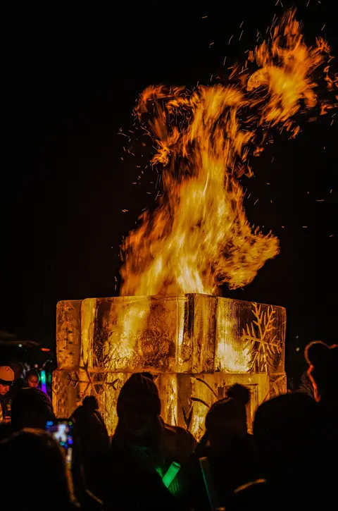 A large bonfire burns brightly inside an ice structure surrounded by people at night.