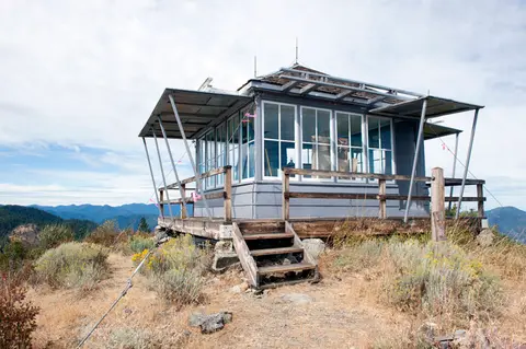 A fire lookout tower stands on a mountaintop surrounded by distant hills and sparse vegetation.