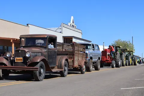 A line of vintage trucks and tractors driving down a town street.