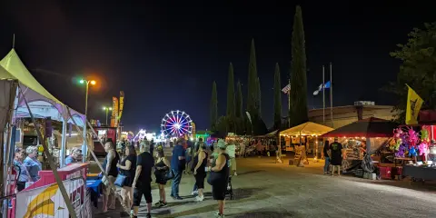 People walk along a lit street fair at night with a Ferris wheel in the distance.