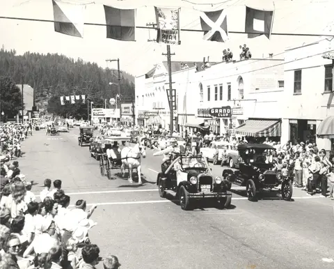 A historic black and white photograph showing a parade on a town street with vintage cars, a horse-drawn carriage, and spectators.