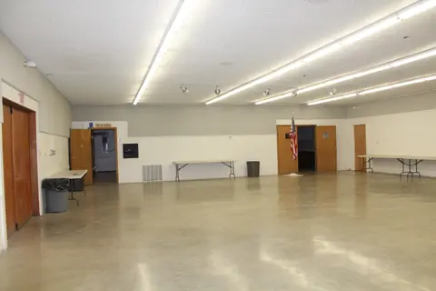 An empty community hall interior with concrete floors and fluorescent lighting.