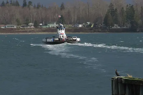 A ferry boat travels across the water with cars on board, and a bird stands on a piling in the foreground.