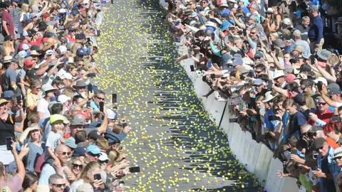 A large crowd of people watches thousands of small yellow balls rolling down a street.