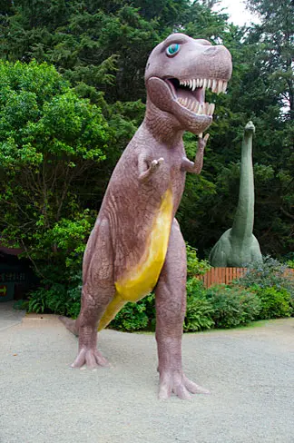 A large dinosaur statue stands outdoors near trees and bushes.