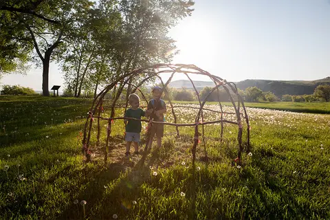 Two children stand inside a willow dome structure in a grassy field.