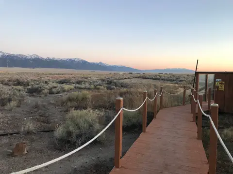 A wooden boardwalk leads toward a building in a landscape with mountains in the background.