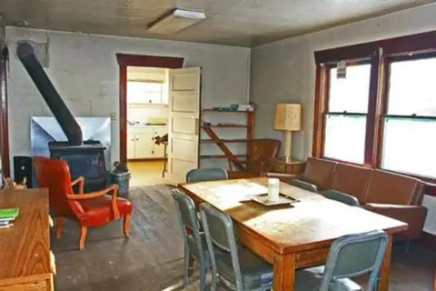 A rustic interior room features a dining table, a wood-burning stove, and a view into another room.