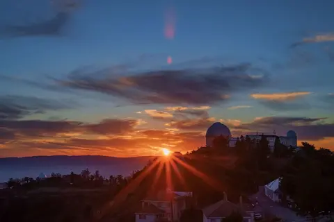 A sunset behind an observatory on a mountain ridge with buildings below.