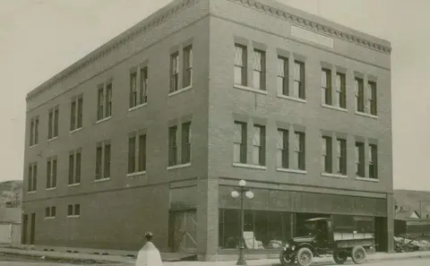 A historic photograph showing a multi-story brick building with a truck parked in front.