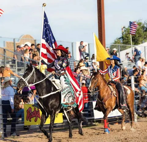 Two women ride horses carrying flags at an outdoor arena event in front of a crowd.