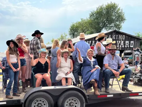 A group of people wearing hats wave from a flatbed trailer outdoors.