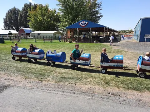 Children ride in a makeshift barrel train outdoors on a sunny day.