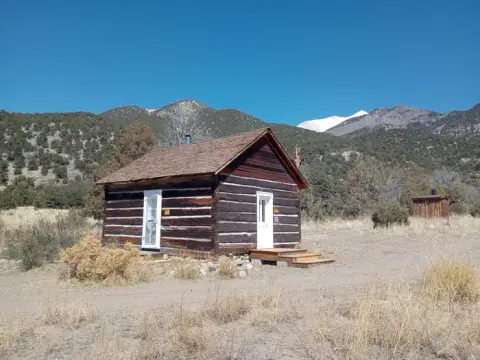 A rustic log cabin sits in a dry landscape with mountains in the background.