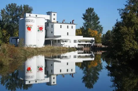 A white flour mill stands by a calm river reflecting its image.