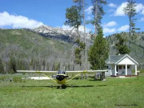 A small airplane parked on a grassy field in front of a cabin and mountains.