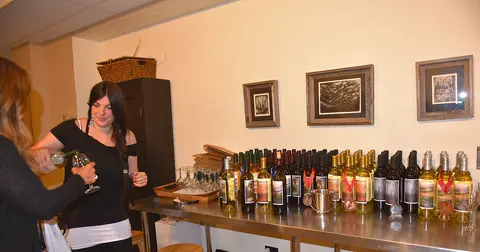 A woman pours wine for another person at a tasting table lined with bottles.