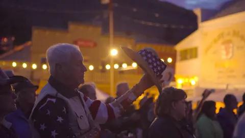 A man holds up a hat with stars and stripes amid a crowd at dusk.