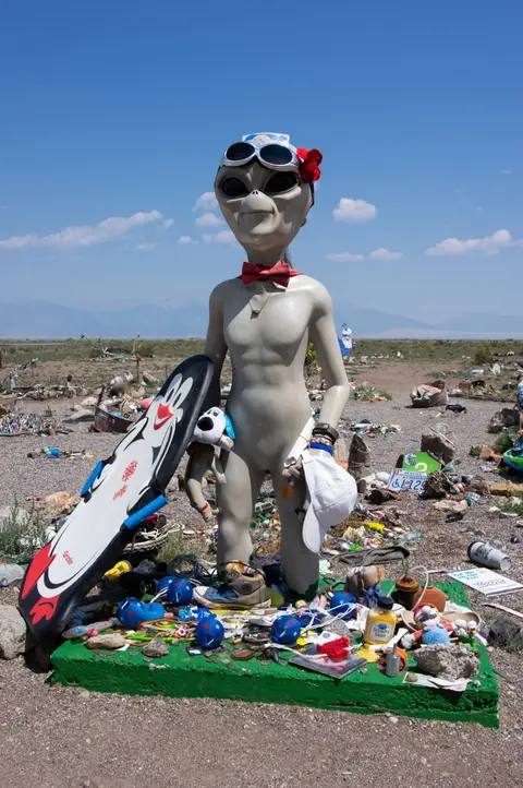An alien statue holding a surfboard stands on a decorated platform outdoors.