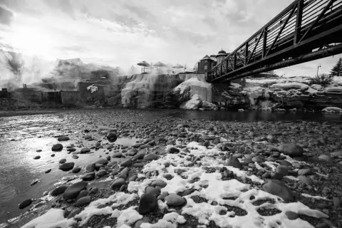 A black and white photo shows a river with a rocky, snow-covered bank, a bridge overhead, and hot springs steaming in the background.