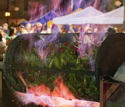 A large metal roaster filled with green chiles sits over flames outdoors with people in the background.