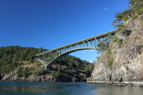 A green suspension bridge spans a body of water between rocky, wooded shores under a clear blue sky.