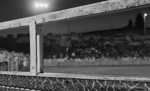 A black and white photo shows a view of an outdoor arena from behind railings at night.