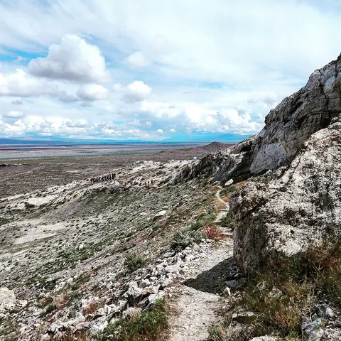 A rocky trail winds down a hillside overlooking a wide valley under a cloudy sky.