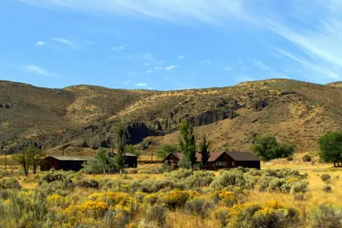 A rustic ranch or homestead sits in a dry landscape below rolling hills under a blue sky.