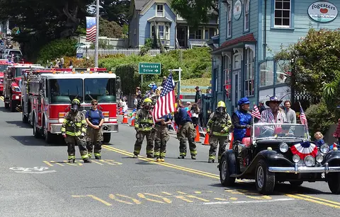Fire trucks and classic cars participate in a parade on a street lined with spectators and buildings.