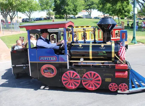 A small replica steam train with passengers being driven outdoors on a street.