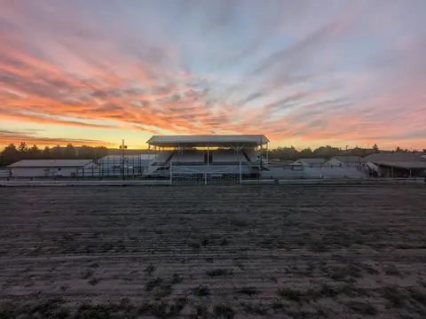 A covered grandstand stands at the edge of a dirt arena under a colorful sunrise sky.