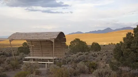 A shaded picnic table stands in a dry, open landscape with mountains in the distance.
