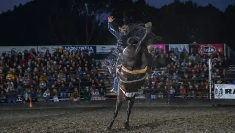 A cowboy rides a bucking horse at a rodeo event in front of a crowd.