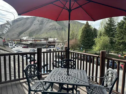A patio set with a red umbrella on a wooden balcony overlooking a street and mountain.