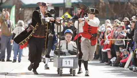People dressed in pirate costumes push a small cart with a rider down a street during an event.