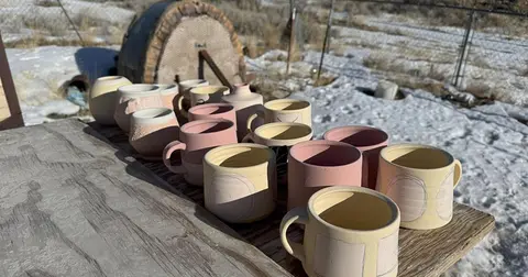 Handmade ceramic cups and pottery sit on a wooden table outdoors with snow visible in the background.