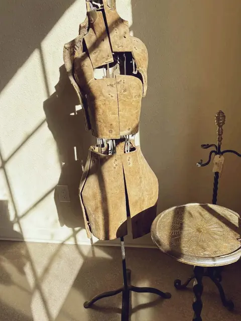 A vintage adjustable dress form stands indoors next to a small ornate table, illuminated by sunlight.