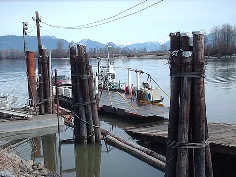 A small ferry docked at a wooden pier on a river with mountains in the background.