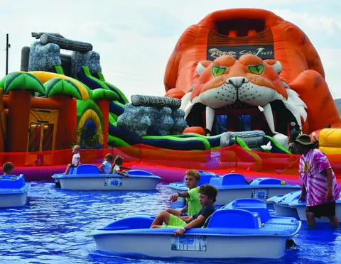 Children ride in small blue paddle boats in a pool in front of large inflatable structures.