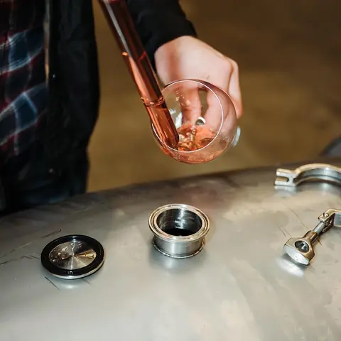 A person uses a wine thief to sample liquid from a metal tank into a glass.