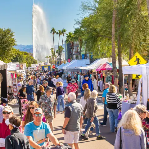 A crowd of people walks along an outdoor market street with a tall fountain visible in the distance.