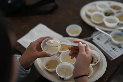 A person holds a small plastic cup and spoon to taste food from a selection of sample cups.
