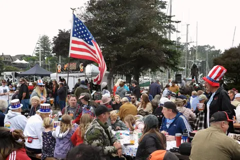 A crowd of people wearing patriotic hats gather outdoors near boats with an American flag flying.