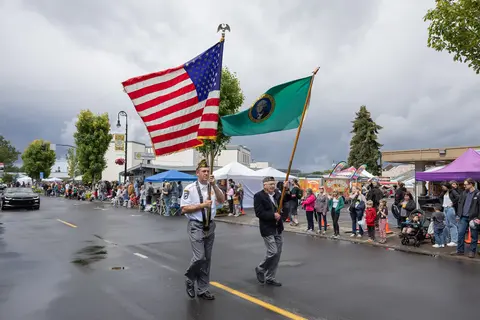 Two veterans carry flags down a wet street during a parade with onlookers on the sidelines.